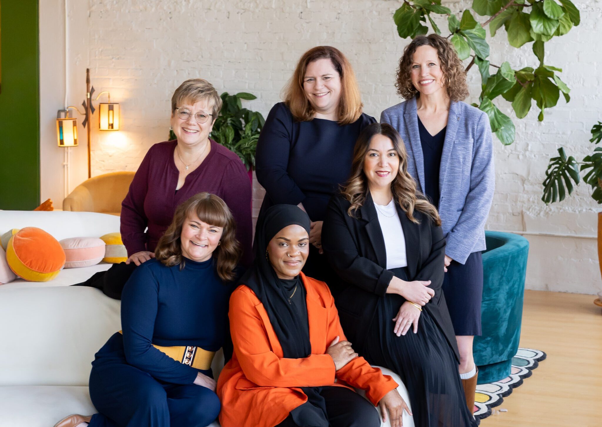 Group portrait of six women in business attire smiling in a contemporary workspace with white brick walls, greenery, and modern furniture.