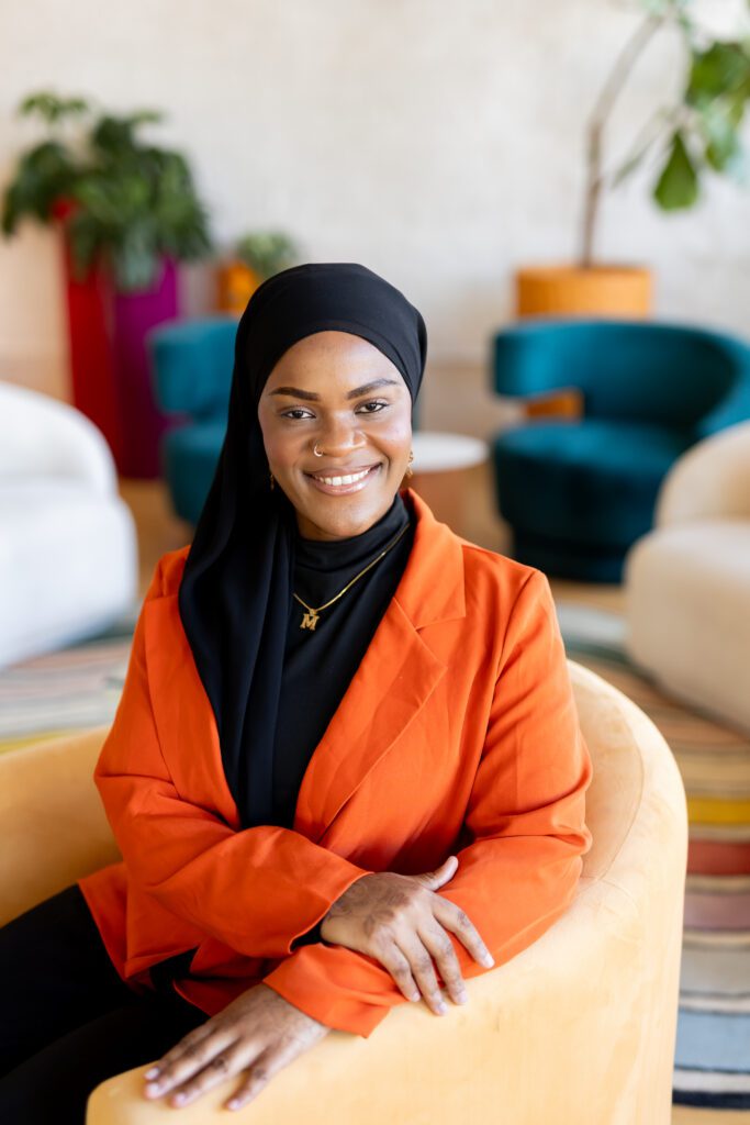 Professional brand photo of a woman in a black hijab and orange blazer seated in a modern office lounge, smiling confidently at the camera.